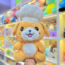 Load image into Gallery viewer, Chef Puppy Soft Toy held in front of a colorful Toy Store Display - Tinyminymo