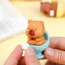 Load image into Gallery viewer, Capybara Eraser on a blue toilet seat with Toilet Paper held by a hand against a blurred background - Tinyminymo

