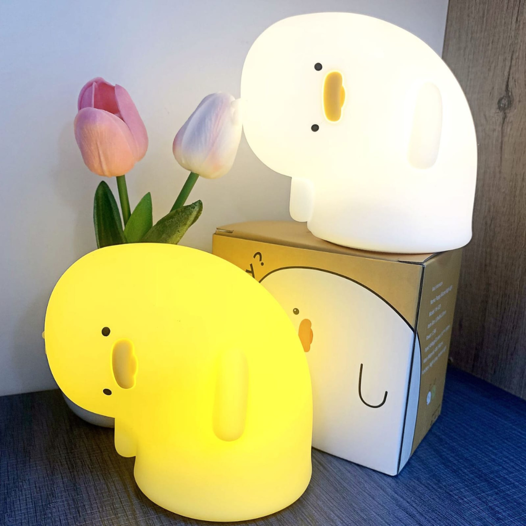 Two Crooked Neck Duck Silicone Night Lights with Floral Decorations in the Background, One Placed on a Box Glowing White, the Other Emitting a Soft Yellow Light - Tinyminymo