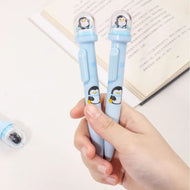 Two Penguin Pens Held in a Hand with a Book in the Background -Tinyminymo