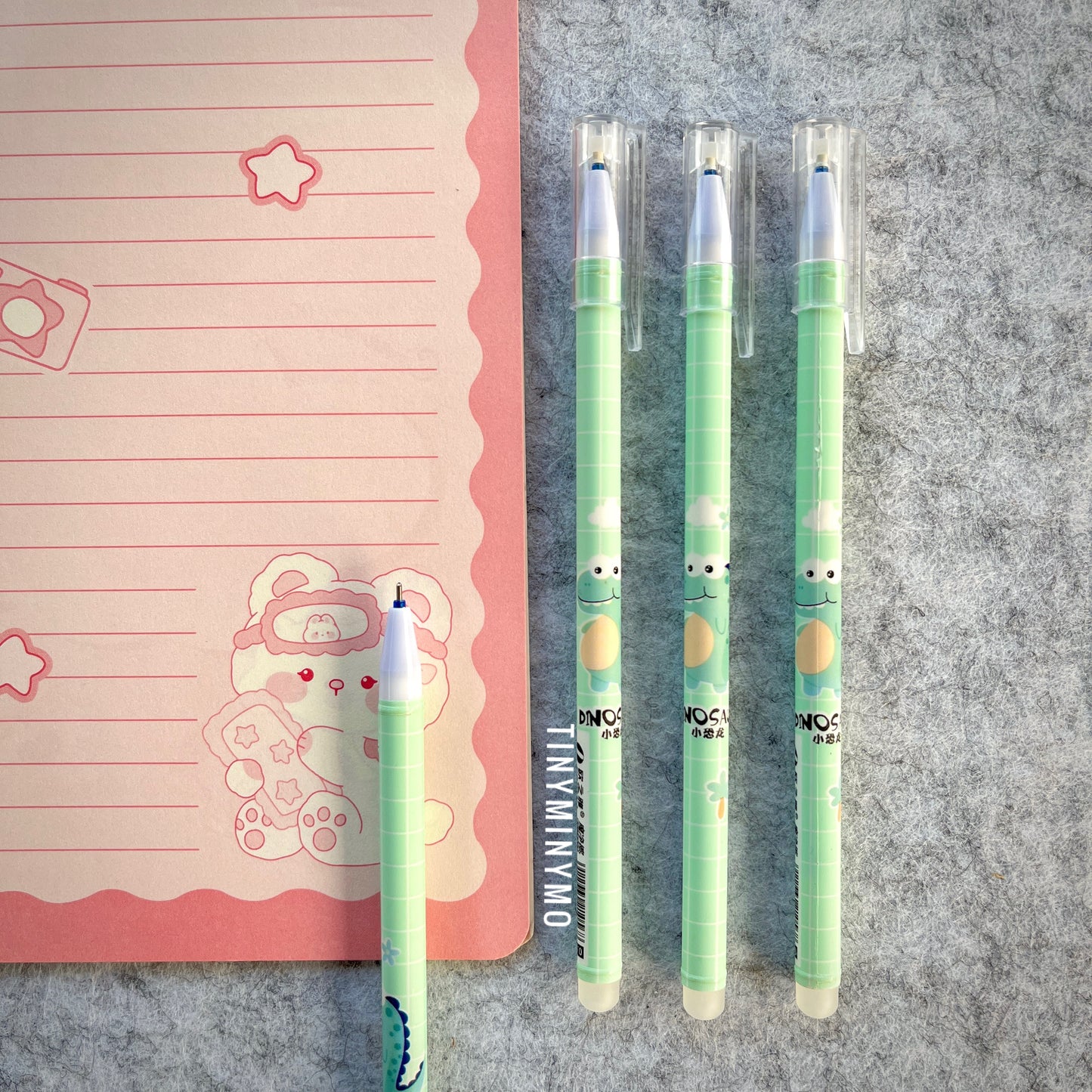 Three Green Pens with Dinosaur Characters on a Grey Surface Next to a Pink Notebook - Tinyminymo