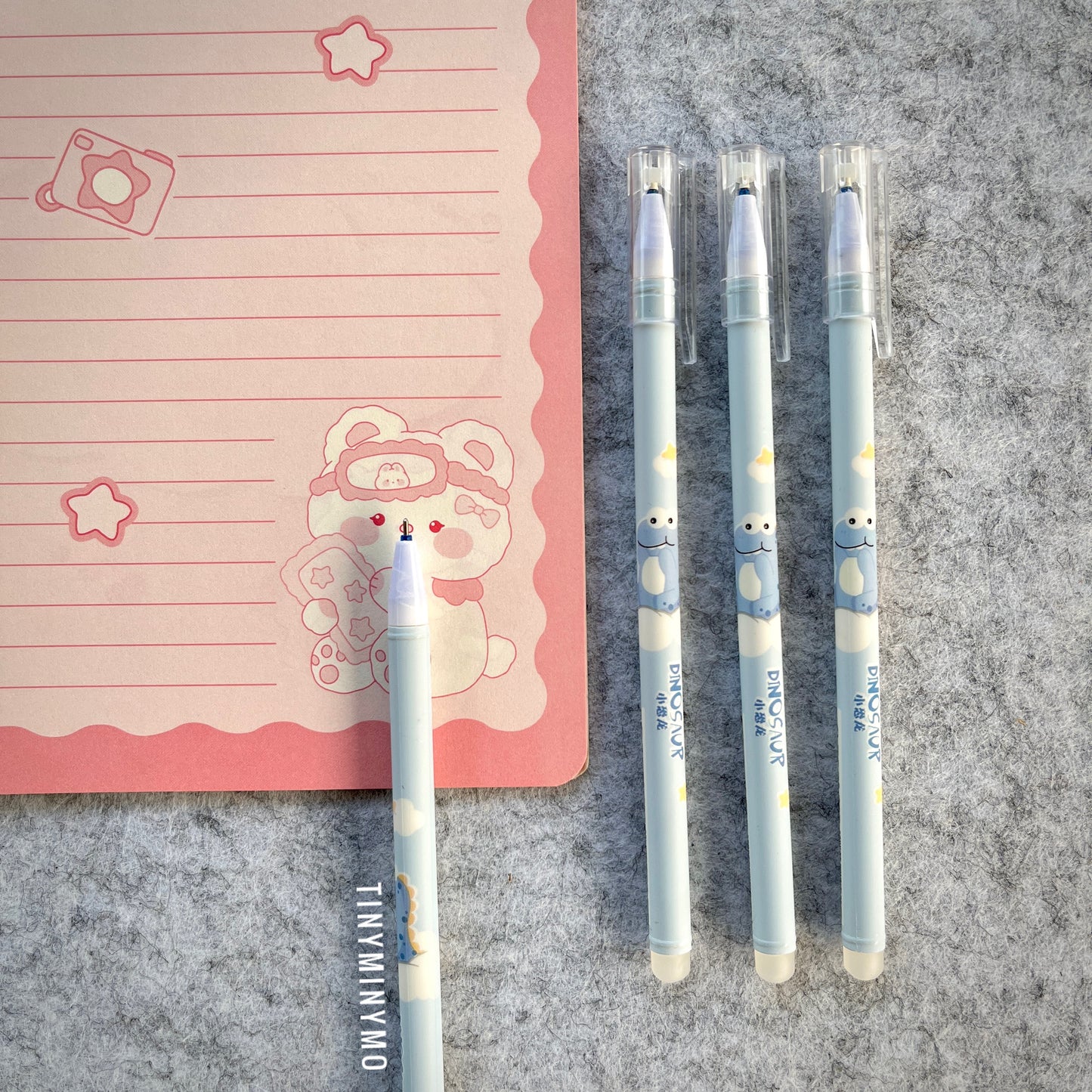 Blue Dinosaur-Themed Pens on a Grey Surface with a Pink Notebook - Tinyminymo