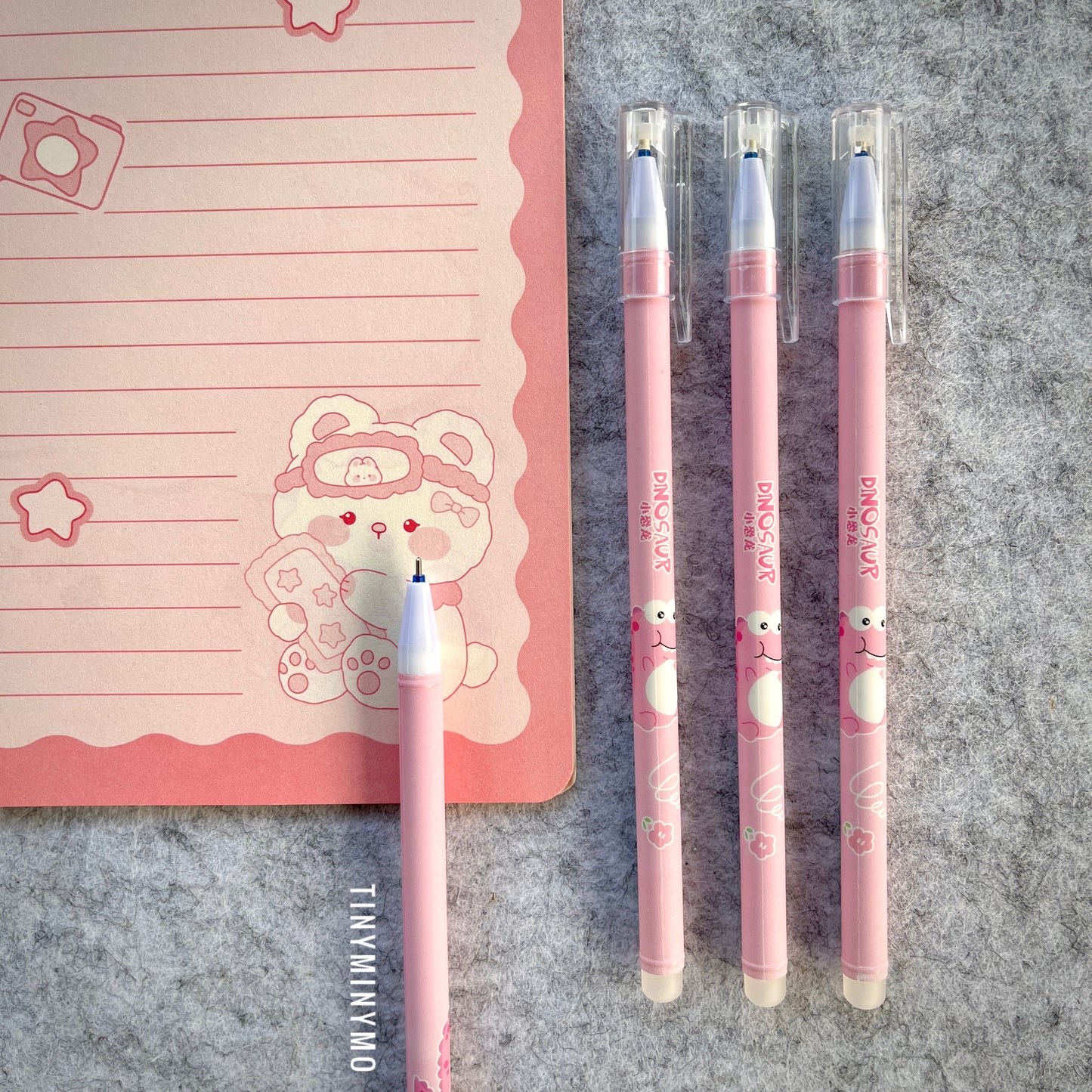 Pink Pens with Dinosaur Design on a Grey Surface Next to a Pink Notebook - Tinyminymo