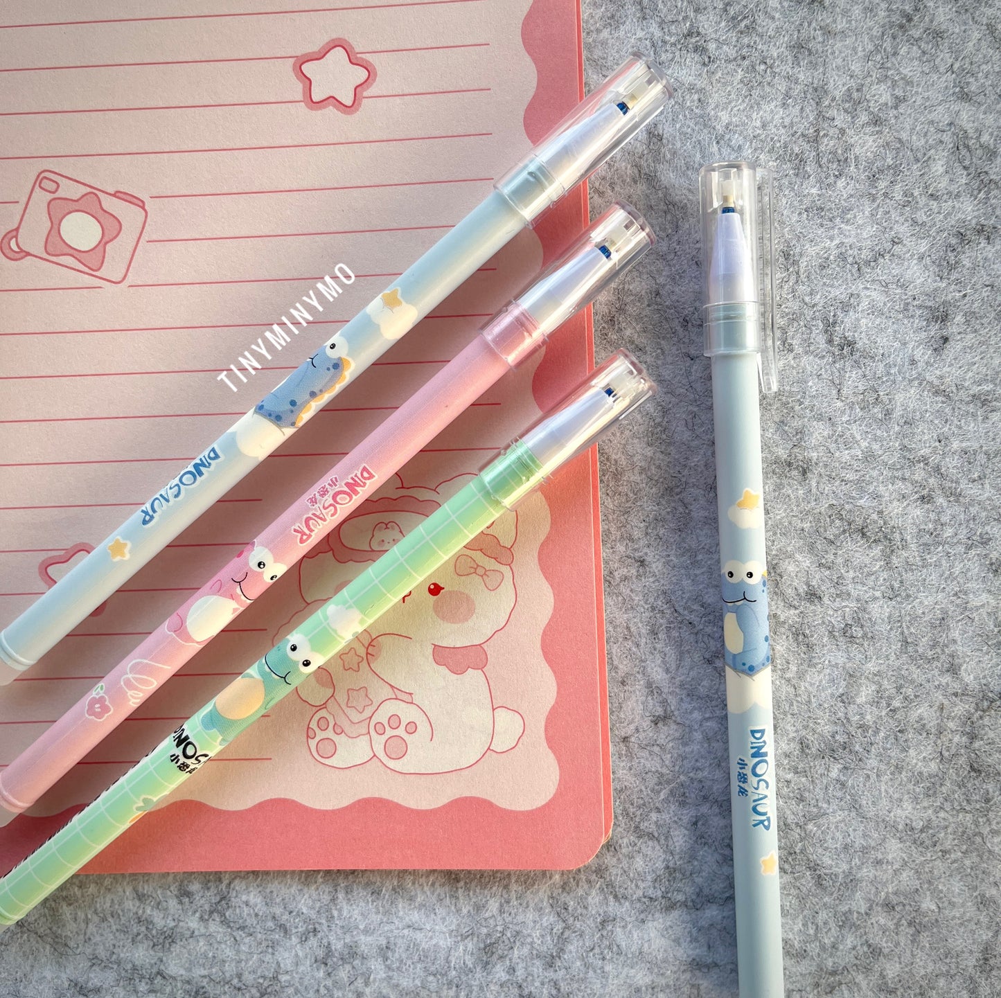 Three Colorful Pens with Dinosaur Designs on a Pink Notebook and Grey Surface - Tinyminymo
