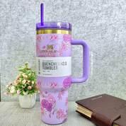 Purple Floral Stanley Cup with a Purple Handle and Straw on a Grey Textured Surface, with a Plant and Notebook in the Background - Tinyminymo