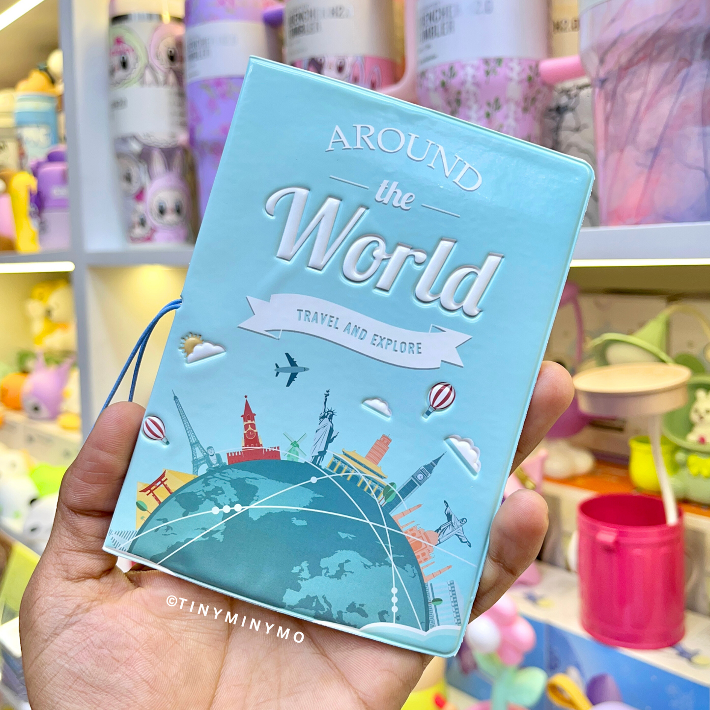 Light Blue Kawaii Passport Cover with 'Around the World' Text, Held in Hand Against a Colorful Background - Tinyminymo