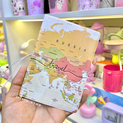 Kawaii Passport Cover with World Map Design Held by a Hand Against a Blurred Background of Colorful Items - Tinyminymo