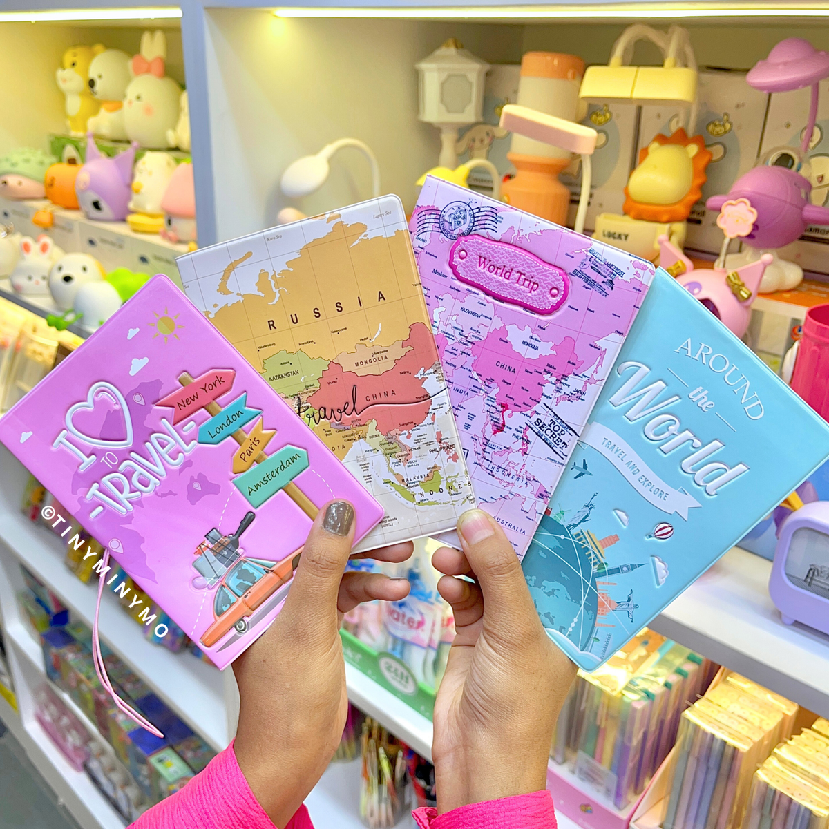 Four Colorful Travel Themed Kawaii Passport Covers Held by a Person in a Store Setting with Toys Visible in the Background - Tinyminymo