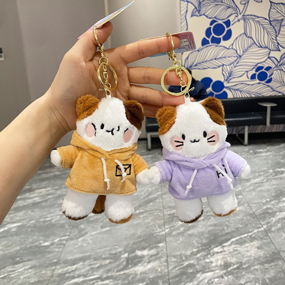 A Hand Holding Two Kitty in Hoodie Plush Keychains Wearing Hoodies in Two Different Colors — Yellow and Purple — Against a Floral Background - Tinyminymo