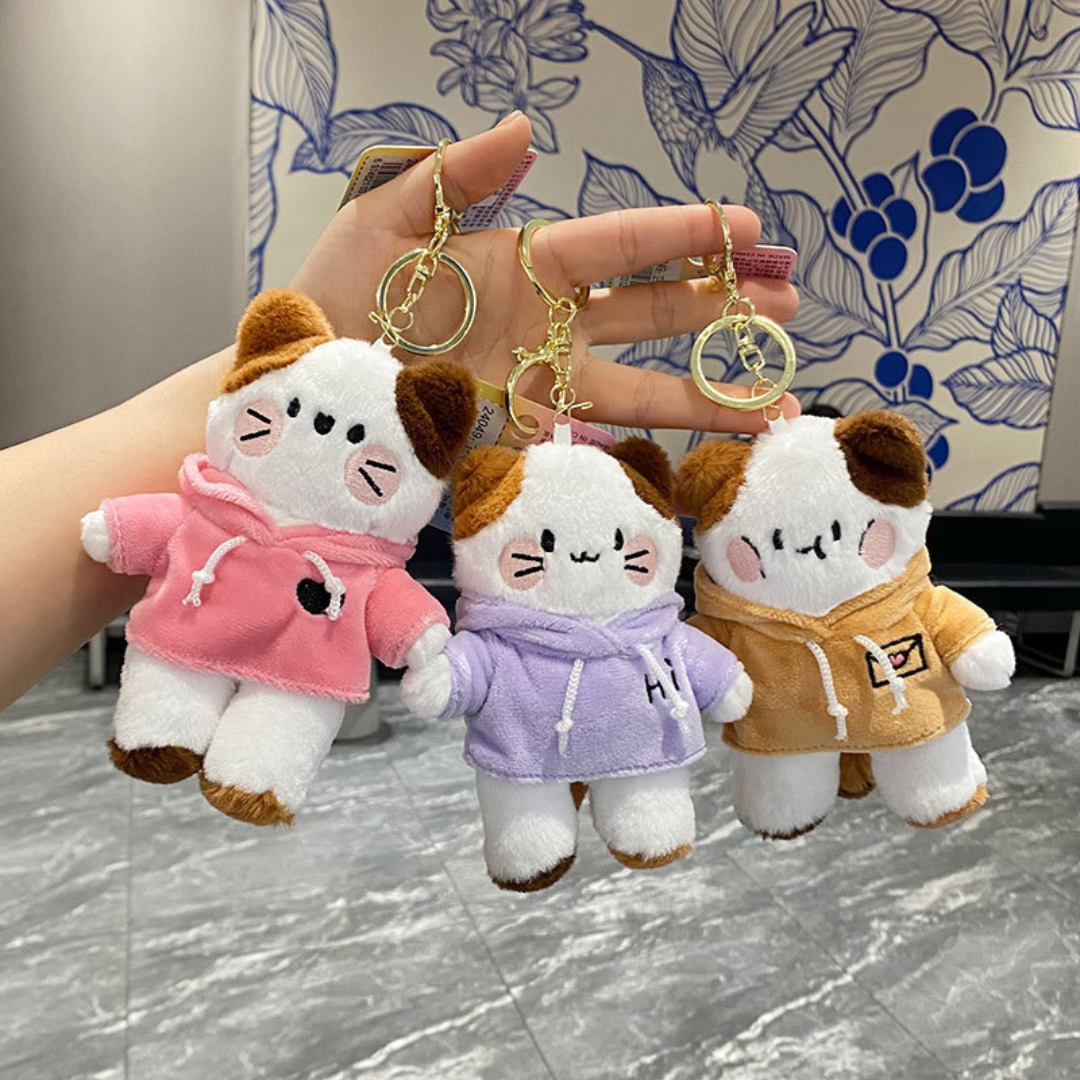 A Hand Holding Three Kitty in Hoodie Plush Keychains in Different Colors — Pink, Purple and Yellow — Against a Floral Wall with a Textured Marble Floor - Tinyminymo