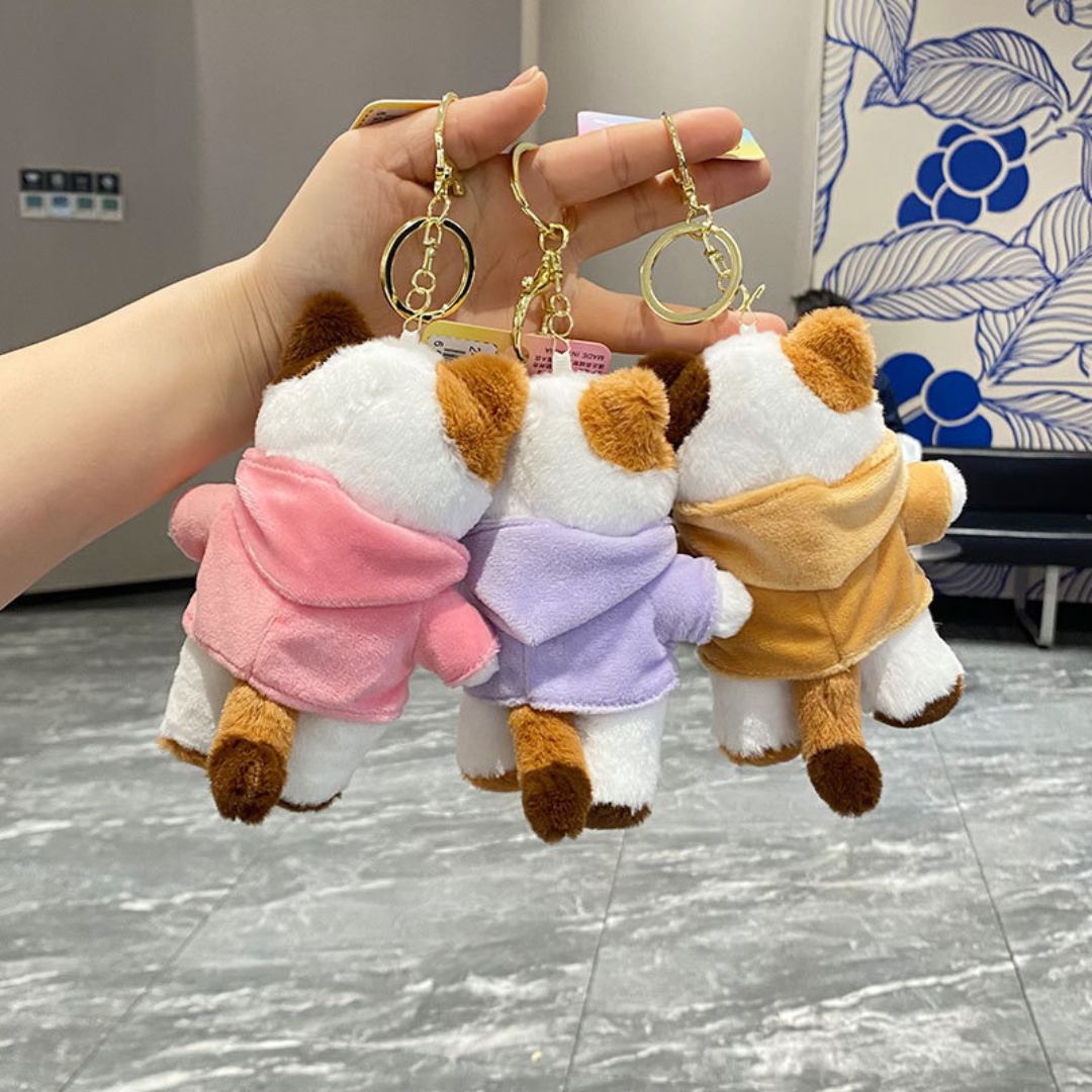 A Hand Holding Three Cute Kitty Plush Keychains Wearing Hoodies in Three Different Colors, Viewed from the Back Against a Floral and Textured Background - Tinyminymo