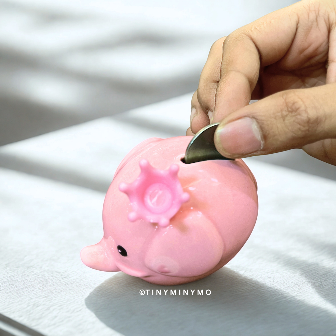 Pink piggy bank with a hand inserting a coin on a light background - Tinyminymo