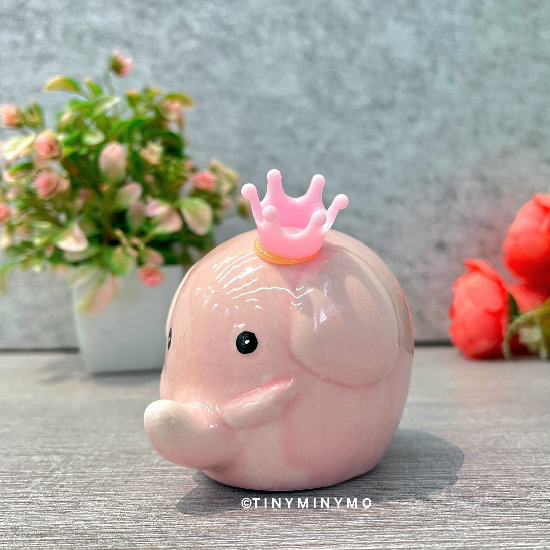 Pink elephant figure with a crown on a gray surface with flowers in the background - Tinyminymo