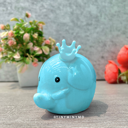 Blue figurine with a crown on a wooden surface with flowers in the background - Tinyminymo