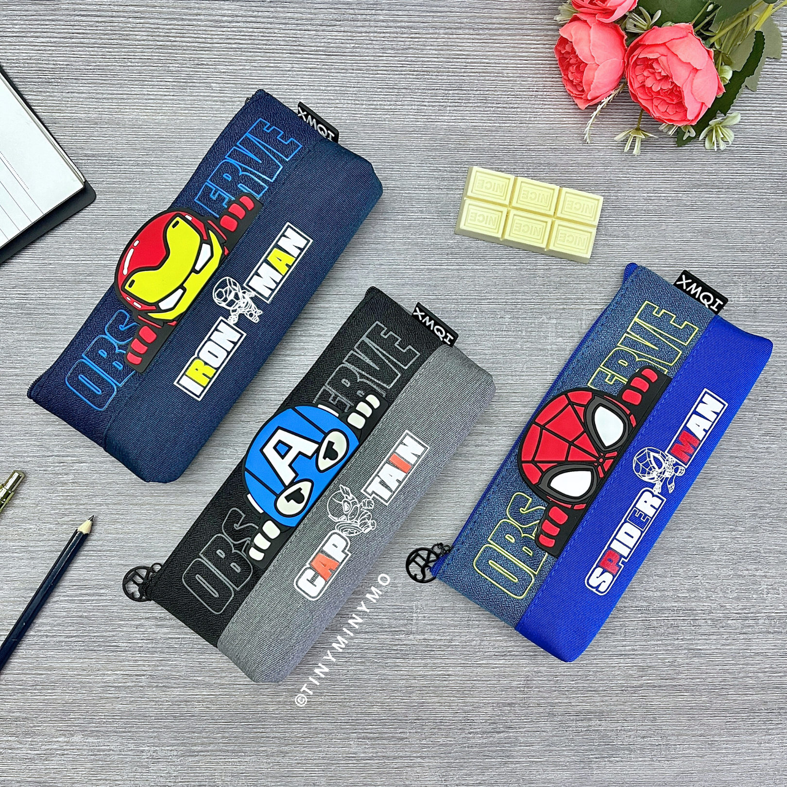 Three Superhero Pencil Pouches with Superhero Designs, Placed on a Grey Surface Alongside a Flowers and a Chocolate Eraser - Tinyminymo
