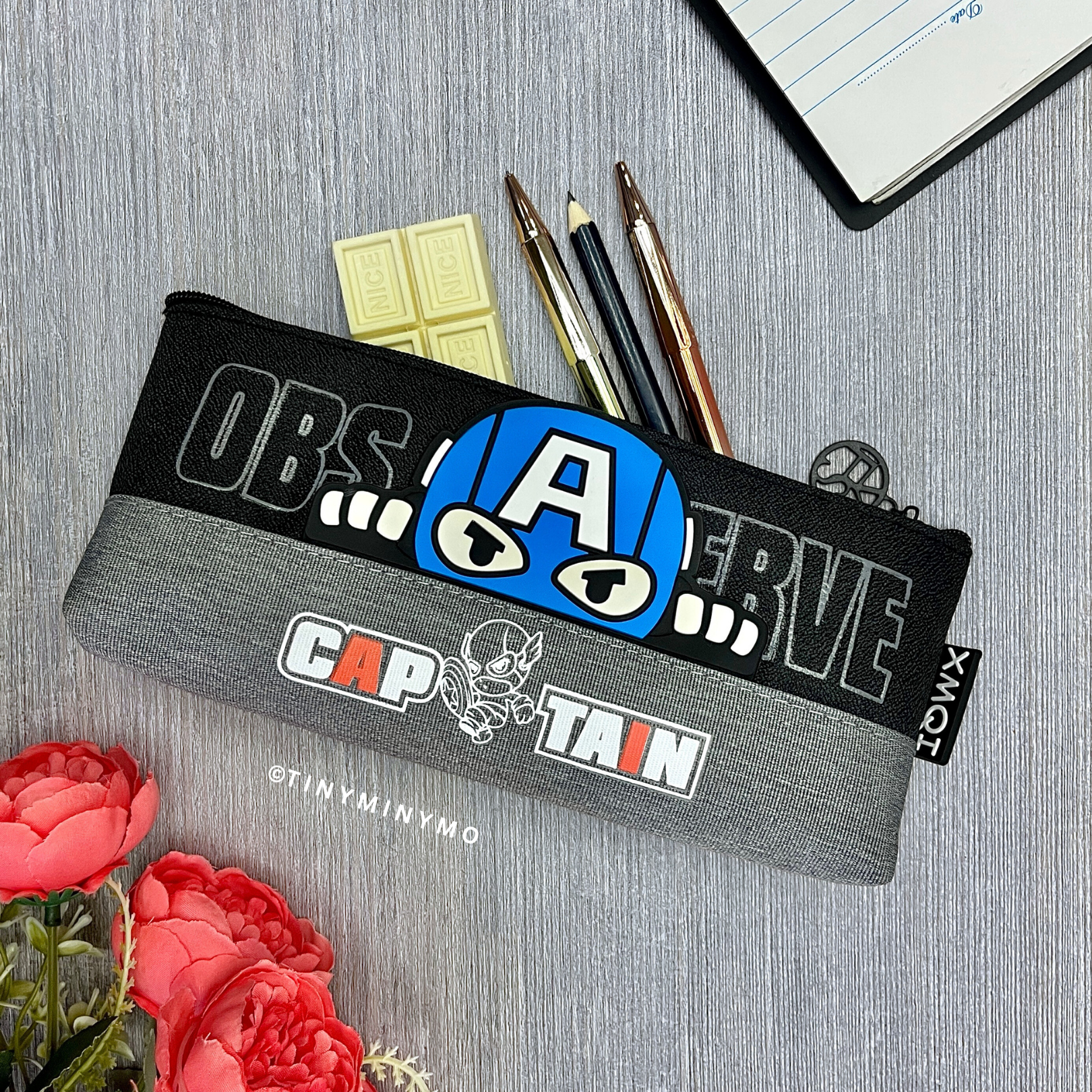 Superhero Pencil Pouch with Various Designs and Text, Placed on a Grey Surface with Flowers and a Notebook - TInyminymo